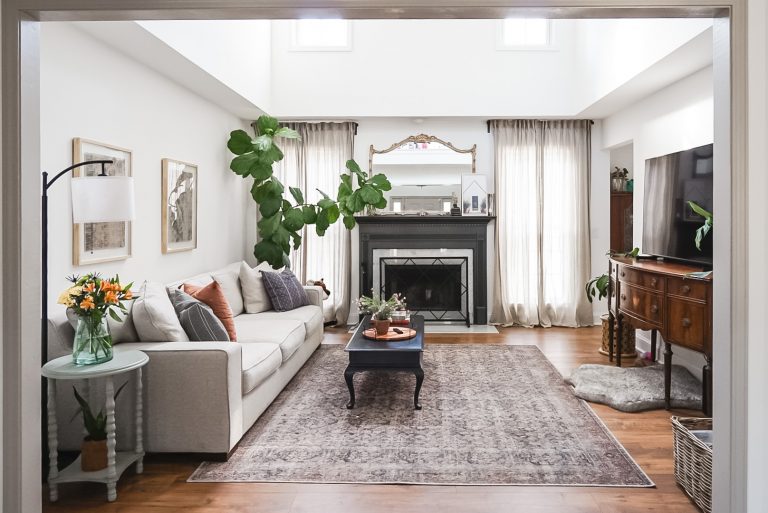 living room with bright natural light, plants, cream colored couch, fireplace with black painted mantle, large rug, TV sitting on antique cabinet
