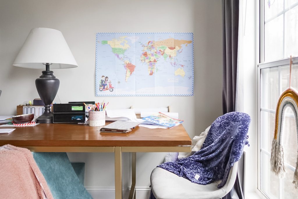 table with homeschool papers and books strewn about, a lamp and pencil holder sit on the table with a map of the world on the wall above table