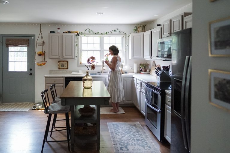 Woman in a dress standing at the kitchen sink with fresh flowers in her hand