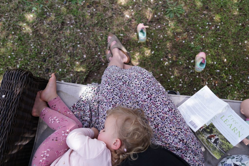 Woman in floral skirt sitting outside reading with toddler napping