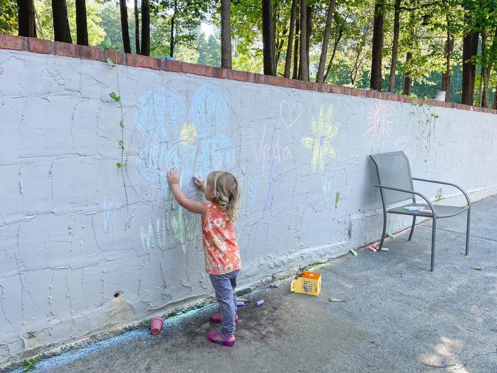 toddler coloring on a retaining wall with chalk and a seat with coffee cup where mom sits