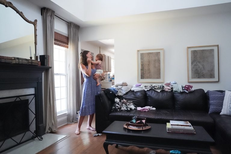 shows woman happily dancing with her baby in living room with laundry piles on couch