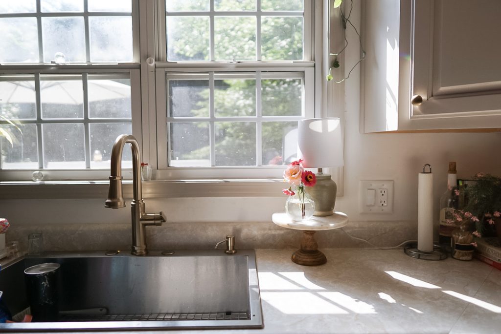 Light pouring through window over kitchen sink. The light lands gently on a clean counter with vase of wildflowers