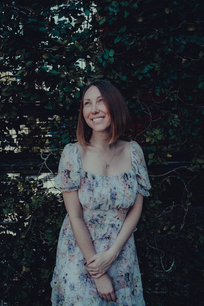 A smiling woman in a floral dress standing outdoors by a lush green hedge.
