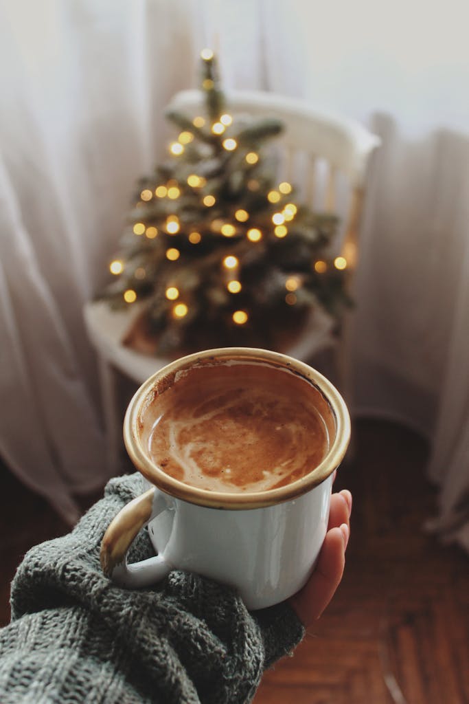 A warm mug of coffee held in hand with a decorated, lit Christmas tree in the background indoors.