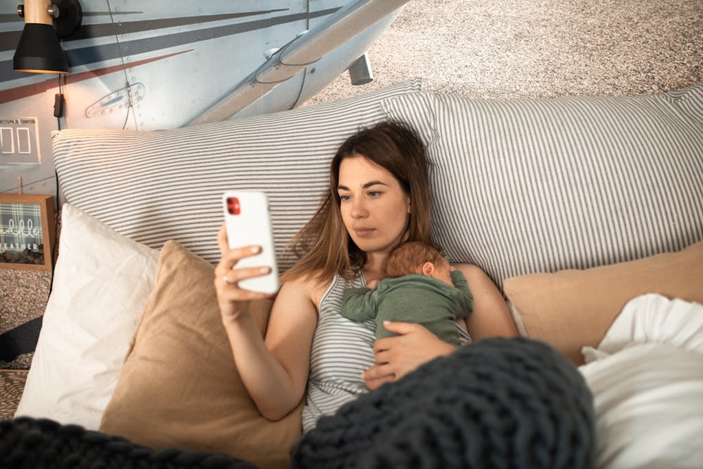 Mother uses smartphone while holding her newborn in bed, surrounded by pillows and cozy decor.