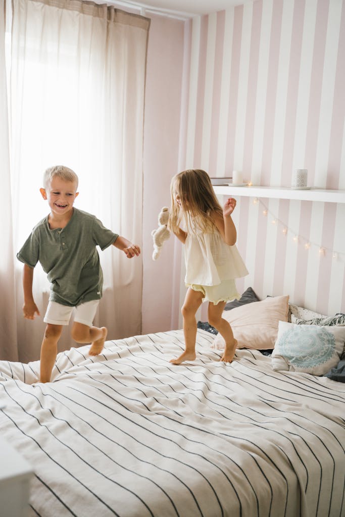 Two kids joyfully jumping on bed in a bright room with soft lighting and playful decor.