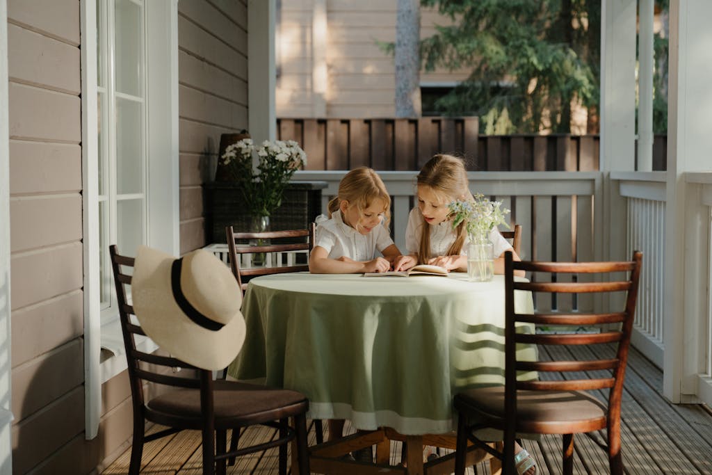 Two young girls sit on a sunlit veranda, enjoying a book together in a peaceful outdoor setting.